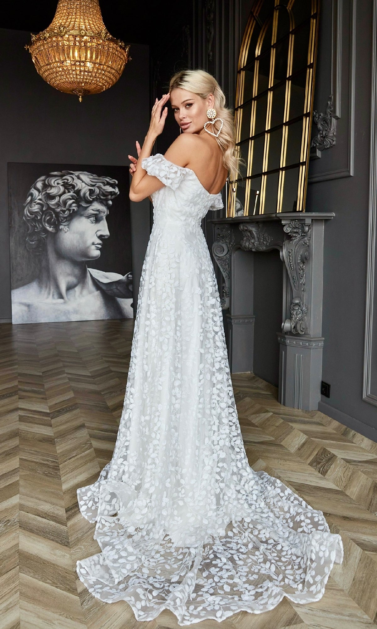 A woman wearing the Tina Holly TK023W long white lace formal dress stands in an elegant room with a chandelier, ornate mirror, and David sculpture artwork.